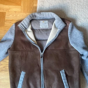 Brun och grå pufferjacka Gran Sasso - Snygg pufferjacka från Gran Sasso med brun framsida i mocka och quiltad grå baksida. Jackan har grå stickade ärmar med bruna mocka armbågslappar, ribbad krage och muddar samt dragkedja framtill. Perfekt för dig som gillar en unik och stilren look. Riktig varm och passar perfekt till kommande årstid, priset är diskuterbart