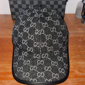 Svart Gucci keps med monogram - UNIK! | Snygg svart keps från Gucci med klassiskt GG-monogram i grått över hela kepsen. Kepsen har en böjd skärm och detaljer i svart läder runt kanten. Tillverkad i bomull med insida i ljus canvas och justerbart spänne bak. Perfekt för dig som gillar lyxiga accessoarer.