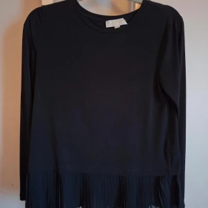 Svart blus med plisserad kant Michael Kors - Svart blus från Michael Kors i storlek M. Toppen har lång ärm och rund hals, med en snygg plisserad kant nedtill som ger extra stil. Materialet är mjuk polyester med lite stretch för skön passform. Perfekt för dig som gillar minimalistisk och chic look.