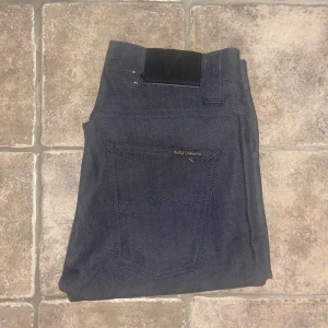 Nudie Jeans Grim Tim  - Snygga mörkblå jeans från Nudie Jeans i modellen Grim Tim. Klassisk femficksdesign, raka ben och normal passform. Tillverkade i 99% bomull och 1% elastan för lite stretch. Midja 36cm, längd 105cm. Ifall det inte finns någon bild i annonsen där jag bär plaggen jag säljer så är det för att jag ej kan ha storleken heller passformen, så kolla måtten för storlek och googla på modellnamnet för passform 😉 s4