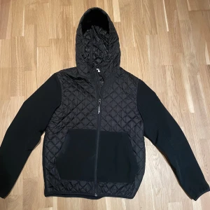 Svart quiltad jacka Tom Tailor Denim - Svart jacka från Tom Tailor Denim i storlek M. Quiltad framsida och huva med snörning, släta svarta ärmar och stor ficka framtill. Dragkedja hela vägen och sportig vibe, perfekt för kyliga dagar.