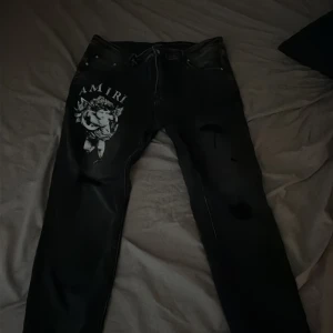 Svarta Amiri jeans med ängeltryck - Snygga svarta jeans från Amiri med stort vitt ängeltryck och logga på vänster lår. Jeansen har klassisk femficksdesign, dragkedja och knapp, samt slitningar på benen för en edgy look. Tillverkade i 100% bomull för skön passform.