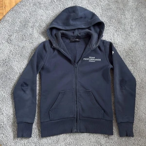 Mörkblå zipup hoodie från Peak Performance - Mörkblå hoodie från Peak Performance med broderad logga på bröstet. Tröjan har dragkedja, huva och två fickor framtill. Insidan är mjuk och fleecefodrad för extra komfort. Perfekt för kyliga dagar och en avslappnad stil. Däremot har armbågarna blivit lite uppslitna, där dragit ner priset enormt. Köpta för 1000, säljs för 200