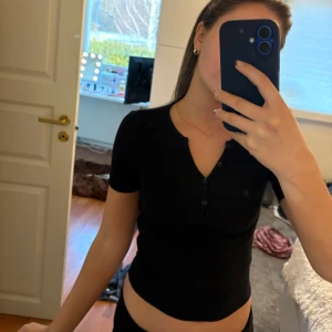 Svart ribbad topp från Gina Tricot - Svart ribbad topp från Gina Tricot i storlek XXS. Modellen har korta ärmar, v-ringad hals med knappar. Passar även XS