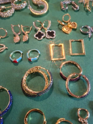 Mix av hoops örhängen i guld och silver - Säljer ett stort urval av örhängen i olika former och färger. Här finns hoops med blå, lila och svarta stenar, hjärtformade hängen, fyrklöver, rektangulära hoops och örhängen med turkos emalj och röda hjärtan. Alla i guld- eller silverfärg, vissa med glittriga detaljer.