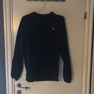 Svart tröja från Lyle & Scott - Svart långärmad tröja från Lyle & Scott med klassisk rund hals och gul broderad fågel på bröstet. Stilren och enkel design som funkar till många olika looks. Materialet känns mjukt och bekvämt mot huden.