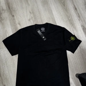 Svart t-shirt från Stone Island - Svart t-shirt från Stone Island med klassisk rund hals och korta ärmar. På vänster ärm sitter den ikoniska Stone Island patchen med broderad logga i gult och grönt. T-shirten är gjord i mjuk bomull och har en clean, stilren look som passar perfekt till streetwear. Ete kvalité 