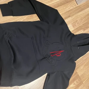 Svart Moncler hoodie med broderad logga - Svart hoodie från Moncler med stor broderad logga i svart och rött på bröstet samt klassisk Moncler-patch på ärmen. Tröjan har huva med metallöljetter och en stor magficka. Perfekt för dig som gillar streetwear och exklusiva märken.