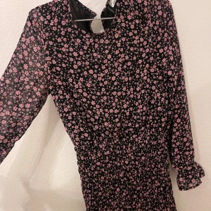 Blommig kortklänning från H&M Divided - Supersöt svart kortklänning från H&M Divided med rosa blommigt mönster. Klänningen har långa ärmar med volangdetalj vid ärmslut och nederkant. Smockad midja ger en snygg passform och tyget är lätt och flowy – perfekt för vår och sommar.