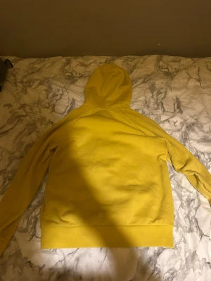 Gul hoodie från Peak Performance - Gul hoodie från Peak Performance med svart logga på bröstet och känguruficka framtill. Tröjan har luva med snörning och ribbade muddar vid ärmslut och nederkant. Perfekt för dig som gillar en chill och sportig stil.