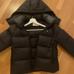 Svart pufferjacka med huva - Svart Calvin Klein vinterjacka, sugen på en ny så känner att jag kan sälja denna. Skitsnygg. Köpte den för 2700kr förra vintern, användes bra men fortfarande som i nytt skick. Priset kan diskuteras 