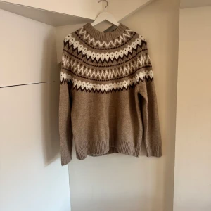 Beige stickad tröja med mönster 157 - Beige stickad tröja från 157 i storlek S med klassiskt nordiskt mönster i vitt och brunt över bröst och axlar. Tröjan har rund halsringning, ribbade muddar och lång ärm. Perfekt för kalla dagar och snygg till jeans eller kjol.