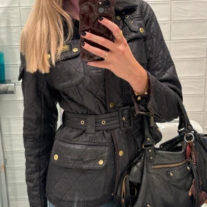 Svart quiltad jacka från Barbour - Quiltad svart jacka från Barbour International med bälte i midjan, guldfärgade knappar och flera fickor. Har använts men är i väldigt bra skick.