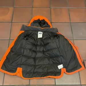 Orange pufferjacka från Helly Hansen - Säljer en fet orange pufferjacka från Helly Hansen med huva och svart innerfoder. Jackan har broderad logga på ärmen och ryggen, samt dragkedja i sidan. Perfekt för kalla dagar när du vill sticka ut med färg och stil. Jag är 181 och den är lite kort. Om du har långa armar är den inte rekommenderad. Pris kan diskuteras🙂