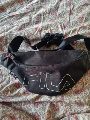 Svart magväska från FILA - Svart magväska från FILA med justerbart band och stort vitt logotryck framtill. Väskan har dragkedja med röd detalj och extra fack på baksidan. Perfekt för att bära det viktigaste och enkel att matcha med streetwear.