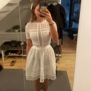 Vit student  spetsklänning  - Superfin vit kort klänning med spets och blommigt mönster. Hög hals, korta ärmar och markerad midja. Klänningen har dragkedja i ryggen och är fodrad under spetsen. Perfekt för student eller sommarens alla tillfällen!