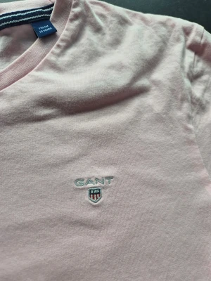 Ljusrosa GANT t-shirt i bomull - Snygg ljusrosa t-shirt från GANT med klassisk rund hals och broderad logga på bröstet. Tillverkad i mjuk bomull som känns skön mot huden. Passformen är normal och ärmarna är korta, perfekt för en chill stil.