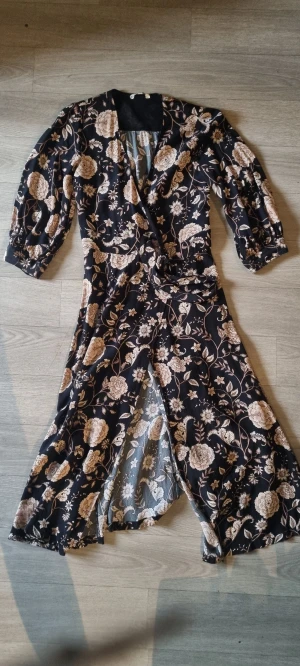 Svart blommig omlottklänning med knyt - Snygg lång omlottklänning i svart med beige blommigt mönster och knyt i midjan. Klänningen har trekvartsärmar med lätt puff och v-ringad hals. Materialet känns mjukt och följsamt, perfekt för dig som gillar en feminin och trendig look.