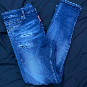 Blå Dsquared2 jeans med slitningar - Snygga blå jeans från Dsquared2 med coola slitningar och distressed detaljer på både fram- och baksida. Klassisk femficksmodell med Dsquared2-logga på röd etikett vid fickan och läderpatch bak. Jeansen har rak passform och är tillverkade i jeansmaterial.