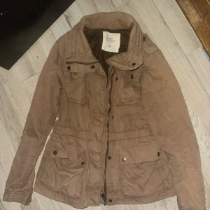 Beige parkajacka från H&M L.O.G.G. - Säljer en beige parkajacka från H&M L.O.G.G. med flera fickor och snygga detaljer som knappar och dragkedja. Jackan har en hög krage och mönstrat innerfoder. Perfekt för dig som gillar en chill och praktisk stil.