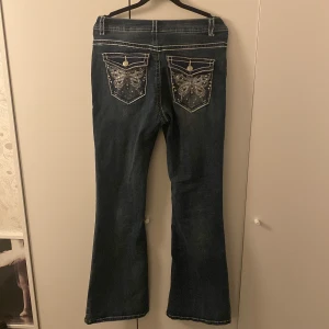 Blå bootcut jeans med broderi från Romwe - Snygga mörkblå bootcut jeans från Romwe med kontrastsömmar och broderade bakfickor med fjärilsmotiv och nitar. Jeansen har klassisk femficksdesign och är tillverkade i bomullsmix för en bekväm passform.