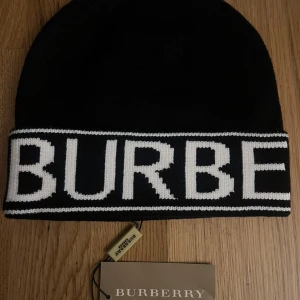 Burberry Mössa - Svart stickad mössa från Burberry med bred vit logotyp runt hela kanten. Klassisk och stilren design som sticker ut med den kontrasterande vita texten. Perfekt accessoar för att lyfta din vinterlook.