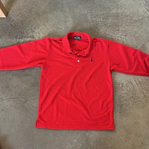 Röd långärmad pikéskjorta Polo Ralph Lauren - Snygg röd långärmad pikétröja från Polo Ralph Lauren med klassisk krage och två knappar framtill. Tröjan är mer röd i verkligheten och har det ikoniska blå broderade Polo-logot på bröstet och är tillverkad i mjuk bomull som är skön mot huden.