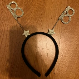 Glittrigt diadem med 18-års tema - Svart diadem med två glittriga siffror '18' i guld på fjädrar och stjärnor. Perfekt accessoar för att fira 18-årsdagen och sticka ut på festen. Diademet är bekvämt och sitter stadigt på huvudet.