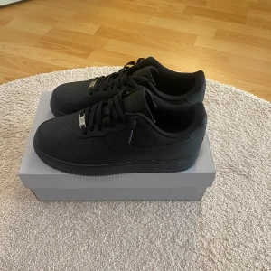 Svarta Nike Air Force 1 sneakers - Säljer ett par helsvarta Nike Air Force 1 sneakers med klassisk låg siluett. Skorna har snörning, perforerad tå och platt sula. Tillverkade i skinn med diskreta detaljer och metallbricka på snörningen. Perfekt för dig som gillar stilrena och tidlösa sneakers.