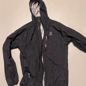 Svart Haglöfs regnjacka med huva - Svart regnjacka från Haglöfs med huva och dragkedja framtill. Jackan har en stilren design med vit logga på bröstet och vattentäta detaljer. Insidan är silverfärgad och materialet är syntet, perfekt för blöta dagar. Långärmad och med praktiska fickor med dragkedja.