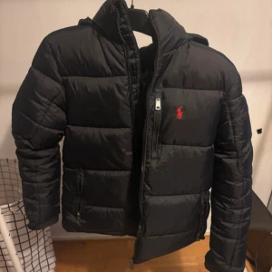Svart pufferjacka från Polo Ralph Lauren - Svart pufferjacka från Polo Ralph Lauren med röd logga på bröstet. Jackan har hög krage, huva och flera dragkedjeförsedda fickor. Den är quiltad och har en clean, sportig look som passar perfekt när det är kallt ute.