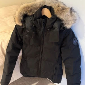Svart dunjacka från Canada Goose - Säljer en svart dunjacka från Canada Goose med fluffig päls på huvan och snygga detaljer som dragkedja och knappar framtill. Jackan har lång ärm, två fickor med dragkedja och patch med logga på ärmen. Perfekt för kalla vinterdagar och riktigt stilren look.