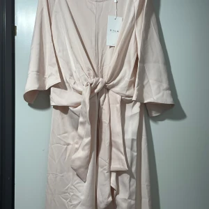 Ljusrosa långklänning från Vila - Superfin ljusrosa långklänning från Vila med knyt i midjan och vida trekvartsärmar. Klänningen har en slits framtill och är tillverkad i ett mjukt, glansigt material som ger en elegant känsla. Perfekt för dig som vill ha en stilren och chic look.