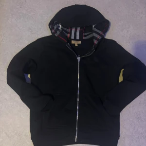 Svart hoodie från Burberry med dragkedja - Svart hoodie från Burberry i storlek M med klassisk rutmönstrad huva i rött, grått och vitt. Tröjan har hel dragkedja framtill, två fickor och är tillverkad i mjuk bomull med lite elastan för extra komfort. Perfekt för dig som gillar stilrena designer med lyxig touch.