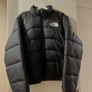 Svart pufferjacka från The North Face - Svart pufferjacka från The North Face med vit logga på bröstet. Jackan har hög krage, lång ärm och dragkedja framtill. Den är quiltad och har en loose passform, perfekt för kalla dagar. Materialet är syntet och dun, vilket gör den både varm och lätt. Endast använd vid ett tillfälle. 