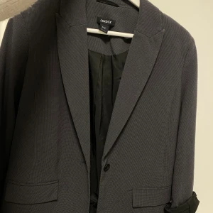 Grå kavaj från Lindex - Snygg grå kavaj från Lindex i diskret rutigt mönster. Klassisk modell med slag, två fickor fram och knäppning med en knapp. Fodrad insida och ärmslut med knappdetalj. 