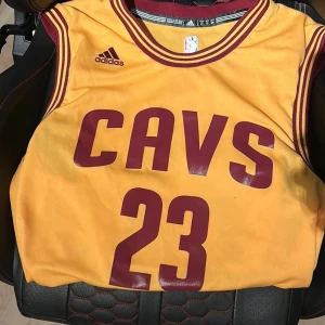 Cleveland Cavaliers James 23 Adidas tröja - Gul baskettröja från Adidas med Cleveland Cavaliers tryck och nummer 23, LeBron James. Tröjan är ärmlös med vinröda detaljer och ribbad krage. Materialet är lätt och ventilerande polyester, perfekt för sport och träning. NBA-logga broderad på ryggen. Pris kan diskuteras 