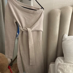 Beige stickad långklänning från NA-KD - Supermysig beige stickad långklänning från NA-KD i storlek XS. Klänningen har offshoulder-design, långa ärmar och ett matchande bälte i midjan som knyts🤍