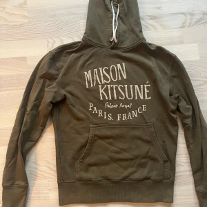 Maison Kitsuné hoodie - Cool olivgrön hoodie från Maison Kitsuné med stor vit text på ryggen. Tröjan har huva med snörning, känguruficka och ribbade muddar. Perfekt för dig som gillar streetwear och fransk design. Materialet är mjuk bomull som känns skönt mot huden.