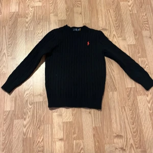 Svart stickad tröja Polo Ralph Lauren - Svart stickad tröja från Polo Ralph Lauren med klassisk kabelstickad design och röd broderad logga på bröstet. Rund hals och långa ärmar, perfekt för dig som gillar stilrena och tidlösa plagg. Skön passform och mjukt material. Och är äkta priset kan vi diskutera privat 