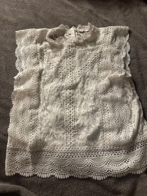 Vit spetsblus från ONLY - Säljer en vit blus från ONLY i storlek S med detaljerad spets och broderade kanter. Blusen har hög hals och är ärmlös med vackra vågiga spetsmönster längs sidorna och nederkanten. Perfekt för dig som gillar romantisk och feminin stil.