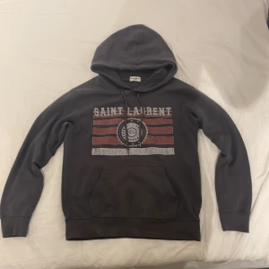 Svart Saint Laurent hoodie med tryck - Svart hoodie från Saint Laurent med huva och snörning. Framsidan har ett coolt tryck med röda och vita ränder samt logga i mitten. Tröjan har en stor magficka och långa ärmar. Perfekt för dig som gillar streetwear och exklusiva märken. Kan också bytas mot en grå likadan fast i M eftersom den är för liten. Skick 9/10 pris kan diskuteras 