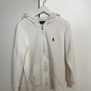 Vit ralph lauren zip hoodie - Vit hoodie från Polo Ralph Lauren med dragkedja och huva. Klassisk design med broderad logga på bröstet, två fickor fram och ribbade muddar. Custom fit-modell i mjuk bomull som är perfekt för chill dagar. Säljer den då jag råkade köpa fel storlek, den är lite mindre i storleken