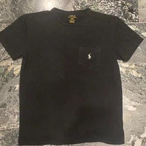 Svart t-shirt från Polo Ralph Lauren - Svart t-shirt från Polo Ralph Lauren med klassisk passform. T-shirten har korta ärmar, rund halsringning och en bröstficka med den ikoniska broderade loggan i vitt. Tillverkad i mjuk bomull för en skön känsla.