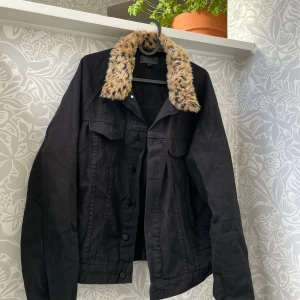 Svart jeansjacka med leopardkrage - Svart jeansjacka med fluffig leopardmönstrad fuskpäls på kragen. Jackan har klassiska bröstfickor med lock och knappar framtill. Perfekt statement-plagg för dig som vill sticka ut med coola detaljer och djurprint.