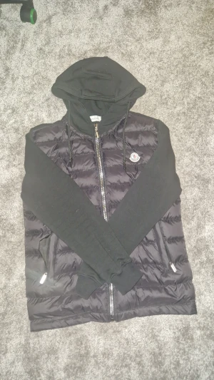 Svart pufferjacka från Moncler L - L i barn size o S i vuxen size. Svart pufferjacka från Moncler med huva och långa ärmar i mjukt tyg. Jackan har quiltad front i glansig polyester, två fickor med dragkedja och Moncler-logga på bröstet. Snygg och chill stil med normal passform och hel dragkedja framtill.