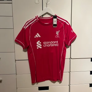 Liverpool FC Salah 11 matchtröja Adidas - Liverpool FCs officiella matchtröja med M. Salah och nummer 11 på ryggen. Röd med vita detaljer, broderat klubbmärke och Adidas-logga. Kortärmad, tillverkad i lätt och ventilerande polyester. Snygga vita ränder på axlarna och tryck från Standard Chartered på bröstet.