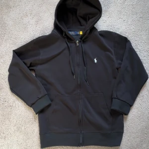 Svart hoodie från Polo Ralph Lauren - Svart hoodie från Polo Ralph Lauren med klassisk vit logobrodering på bröstet. Tröjan är lite liten i storleken så skulle säga att den är S/XS (ny med prislapp)