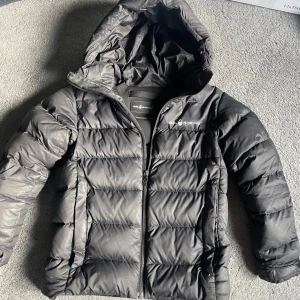 Svart pufferjacka från Sail Racing - Svart pufferjacka från Sail Racing med huva och dragkedja framtill. Nypris 3000 och passar från 150 till 165 cm