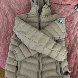 Beige pufferjacka från Vinson Polo Club - Säljer en beige pufferjacka från Vinson Polo Club i storlek S. Jackan har huva, dragkedja framtill och två sidofickor med dragkedja. Puffad design med snyggt märke på ärmen. Perfekt för kyliga dagar och har en clean look.